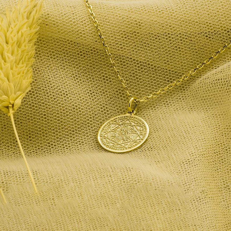 May include: A gold necklace with a round pendant featuring a detailed geometric design. The necklace is displayed on a yellow fabric background.