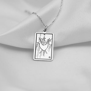 Puede incluir: Collar de plata con un colgante rectangular que presenta una ilustraci&oacute;n en blanco y negro de la carta del tarot Tres de Espadas. La carta representa tres espadas que atraviesan un coraz&oacute;n.