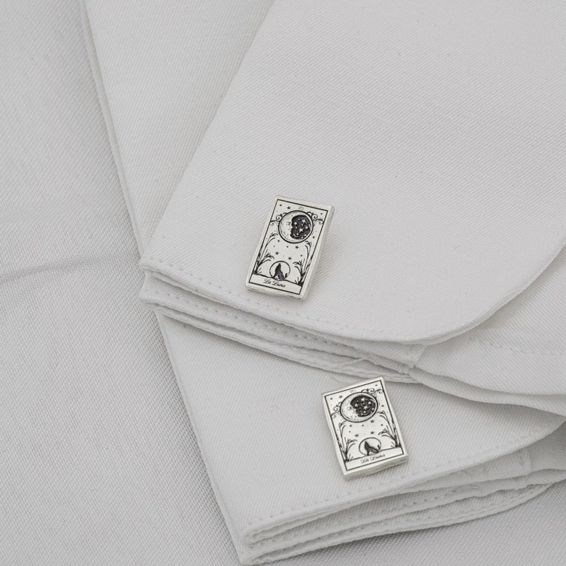 May include: Silver cufflinks with a rectangular design featuring a crescent moon, stars, and a howling wolf. The cufflinks are set against a white background, showcasing the intricate details of the celestial and animal imagery.