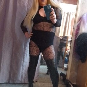 May include: A person wearing a black bodysuit with a spiderweb pattern and black thigh-high boots. The bodysuit has a lace neckline and the boots have a pointed toe.