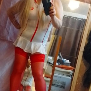 Puede incluir: Una mujer con un disfraz de enfermera blanco con detalles rojos, medias rojas y tacones blancos. Está sosteniendo un teléfono inteligente negro y mirando a la cámara.