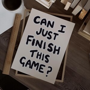 May include: A framed print with the text "Can I just finish this game?" written in black marker on a white background.