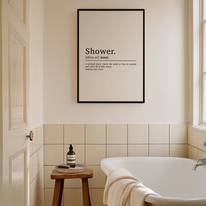 May include: A black framed print with the word "Shower" in white text and a definition of the word. The print is hanging on a white tiled wall in a bathroom with a white bathtub.