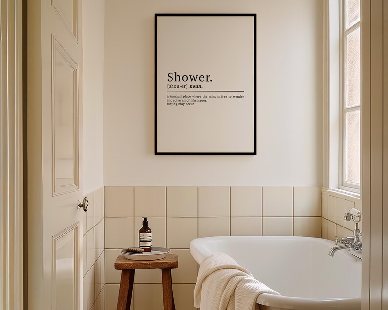 May include: A black framed print with the word "Shower" in white text and a definition of the word. The print is hanging on a white tiled wall in a bathroom with a white bathtub.