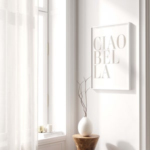 May include: A white framed print with the text "CIAO BELLA" in a light brown color. The print is hanging on a white wall next to a window with white sheer curtains. A white vase with brown branches is on a wooden stool.