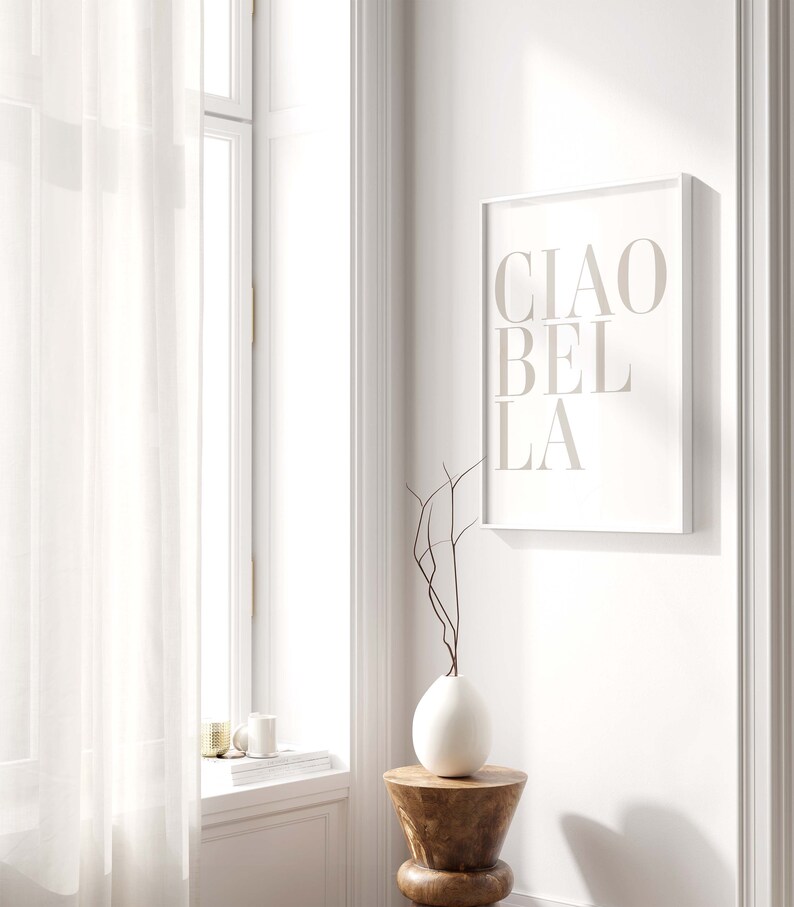 May include: A white framed print with the text "CIAO BELLA" in a light brown color. The print is hanging on a white wall next to a window with white sheer curtains. A white vase with brown branches is on a wooden stool.