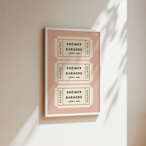 May include: A framed print featuring three vintage-style tickets. Each ticket reads "SHOWER KARAOKE ADMIT ONE" in black text on a cream background, with a pink border. The tickets are arranged vertically on a pink background.