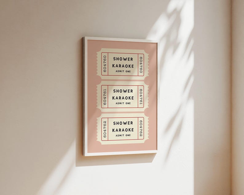 May include: A framed print featuring three vintage-style tickets. Each ticket reads "SHOWER KARAOKE ADMIT ONE" in black text on a cream background, with a pink border. The tickets are arranged vertically on a pink background.