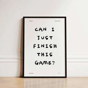 May include: A framed black and white print featuring the text "CAN I JUST FINISH THIS GAME?" in a bold font. The print is part of the "GAMERS COLLECTION" and is displayed on a light-colored wall and wooden floor.