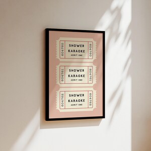 May include: A framed print featuring three vintage-style tickets. Each ticket reads "SHOWER KARAOKE ADMIT ONE" in black text on a cream background, with a pink border. The frame is black, and the background is a light pink.