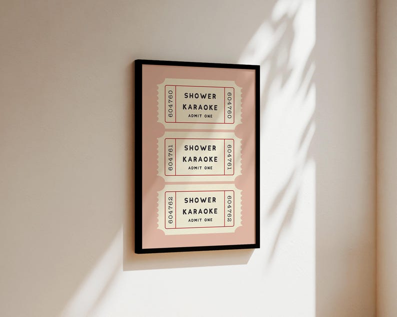 May include: A framed print featuring three vintage-style tickets. Each ticket reads "SHOWER KARAOKE ADMIT ONE" in black text on a cream background, with a pink border. The frame is black, and the background is a light pink.