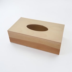 Könnte beinhalten: Eine rechteckige Taschentuchbox aus Holz mit einer hellen, glatten Oberseite und einer dunkleren Seitenwand mit sichtbarer Holzmaserung. Die Oberseite verfügt über eine mittig platzierte, längliche ovale Öffnung zur Entnahme.