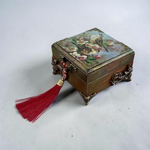 Könnte beinhalten: Kleine Holzbox mit Scharnierdeckel, verziert mit einem Gemälde von Vögeln und Blumen. Die Box hat Eckdetails und Füße aus Metall. Eine rote Quaste hängt von der Vorderseite. Die Box ist etwa 10 cm lang.