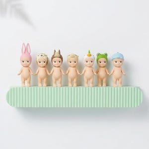 May include: Seven small, nude-colored figurines with various head coverings, including bunny ears, a chef's hat, and a frog hat, stand on a light green, ribbed shelf. The shelf is against a white background.