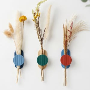 May include: Three wall-mounted vases with dried flowers. Each vase has a wooden base, a coloured circle, and a blue, green, or red accent. The vases hold various dried grasses and flowers, creating a minimalist, modern aesthetic.