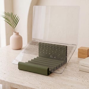 Op de afbeelding: Een doorzichtige acryl laptopstandaard met een donker olijfgroene siliconen basis voorzien van een reeks horizontale ribbels. De standaard staat in een hoek om een laptop te ondersteunen, waarbij het toetsenbord door het materiaal zichtbaar is.