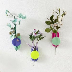 May include: Three wall-mounted, colourful, geometric vases with flowers. Each vase has a different colour scheme: purple and green, purple and yellow, and pink and green. A small vase with a two-tone design and a geometric sculpture are on a shelf.