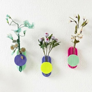May include: Three colorful wall-mounted vases with faux flowers. The vases are in shades of green, blue, and pink, each with a contrasting circular accent. The vases hold different types of artificial flowers, creating a vibrant and modern display.