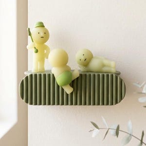 May include: A decorative shelf with three whimsical figures. The figures are in shades of yellow and green, with one holding a stick. The shelf is olive green and has a ribbed design. The figures are on a white wall.