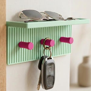 Puede incluir: Un estante organizador de pared de color verde claro con textura acanalada vertical y tres ganchos cilíndricos de color rosa brillante. El estante sostiene unas gafas de sol oscuras, mientras que un llavero de metal con una llave de coche negra cuelga del gancho central.