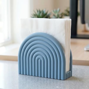 May include: A blue napkin holder in a rainbow design holds a stack of white napkins. The holder is made of a solid material and sits on a light-colored surface. The design is a modern, decorative piece for the home or office.
