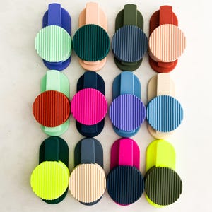 May include: A selection of vibrant phone grips in a range of colours, such as green, pink, blue, and yellow. Each grip has a circular, ridged design and a rectangular base for attaching to a phone or device.