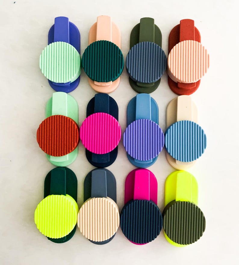 May include: A selection of vibrant phone grips in a range of colours, such as green, pink, blue, and yellow. Each grip has a circular, ridged design and a rectangular base for attaching to a phone or device.
