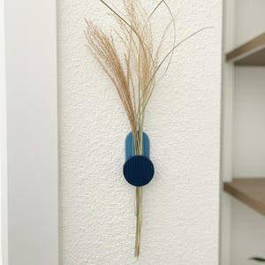 May include: A blue wall-mounted vase with dried ornamental grass. The vase features a circular accent and a rounded top. The wall is white with a textured finish. A shelf is visible in the background.
