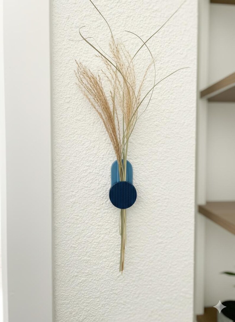 May include: A blue wall-mounted vase with dried ornamental grass. The vase features a circular accent and a rounded top. The wall is white with a textured finish. A shelf is visible in the background.
