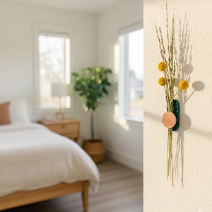 May include: A teal and copper-coloured wall vase holding dried yellow flowers and grasses. The vase is mounted on a textured, off-white wall. The background shows a bedroom with a bed and a potted plant.