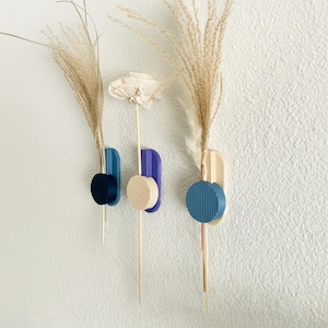 May include: Three wall-mounted vases in blue, beige, and purple, each holding dried floral stems. The vases have a geometric design with a circular accent. The wall is white.