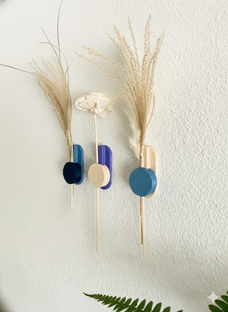 May include: Three wall-mounted vases in blue, beige, and purple, each holding dried floral stems. The vases have a geometric design with a circular accent. The wall is white.