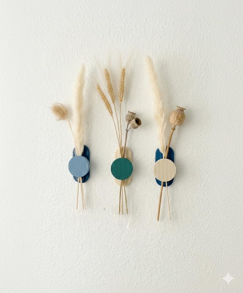 May include: Three wall-mounted vases with dried flowers. Each vase has a dark blue, light blue, and beige colour scheme with a circular design. The vases hold wheat, pampas grass, and seed pods.