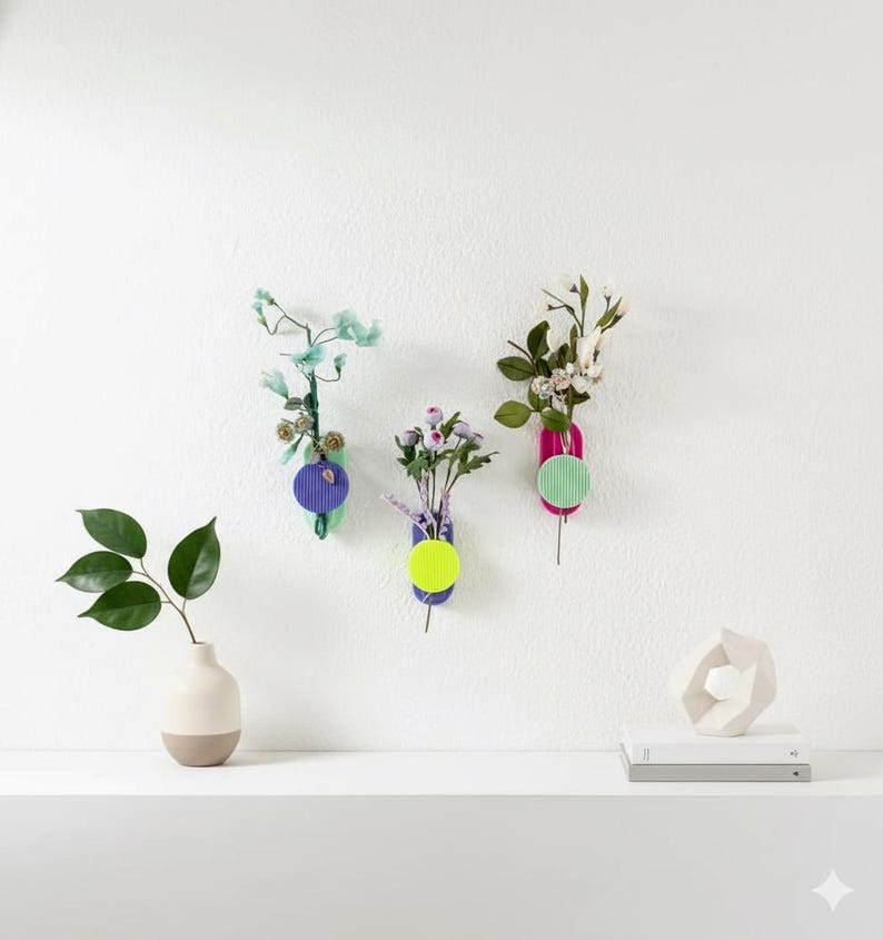 May include: Three wall-mounted, colourful, geometric vases with flowers. Each vase has a different colour scheme: purple and green, purple and yellow, and pink and green. A small vase with a two-tone design and a geometric sculpture are on a shelf.