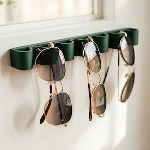 May include: A dark green wall-mounted organizer featuring a wavy, undulating design with multiple slots. Three pairs of sunglasses with thin metal frames and dark lenses are hanging from the individual curved sections of the holder.