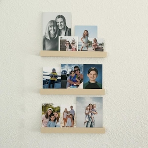 May include: Three wooden floating shelves mounted on a white wall, each holding multiple framed photographs. The photos depict various family portraits and candid shots, showcasing a range of people and settings. The shelves are a light wood color.