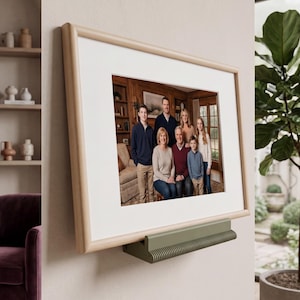 May include: A rectangular picture frame with a light wood finish and a white mat, displaying a family portrait of seven people. The frame is mounted on a wall and rests on a horizontal, olive green, ribbed metal ledge or shelf support.