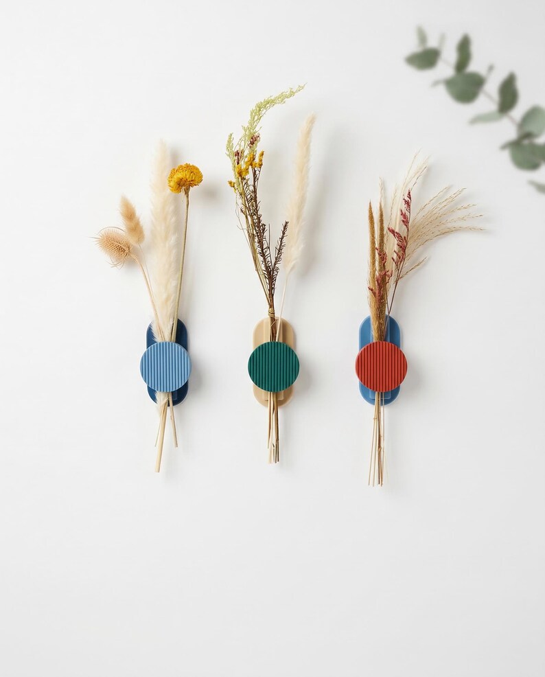 May include: Three wall-mounted vases with dried flowers. Each vase has a wooden base, a coloured circle, and a blue, green, or red accent. The vases hold various dried grasses and flowers, creating a minimalist, modern aesthetic.