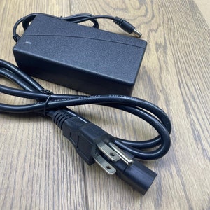 Puede incluir: Adaptador de corriente negro con un cable de alimentación negro y un enchufe español. El adaptador tiene el texto "SMPS7-03K  A005/800V  3X0.75MM2  1A/40V" impreso en él.