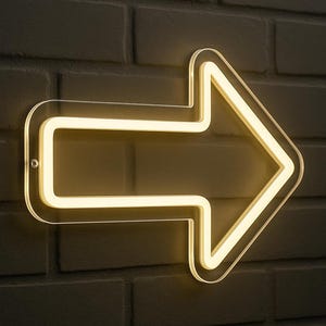 Könnte beinhalten: Ein Neonpfeil-Schild mit klarer Acrylkante, montiert an einer dunklen Backsteinmauer. Der Pfeil leuchtet mit warmem, gelbem Licht nach rechts. Geeignet für Heimdekoration oder gewerbliche Nutzung.