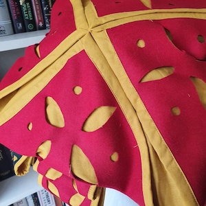 Puede incluir: Un sombrero de tela roja con ribete dorado y dise&ntilde;os recortados. El sombrero est&aacute; doblado y parece ser de fieltro.
