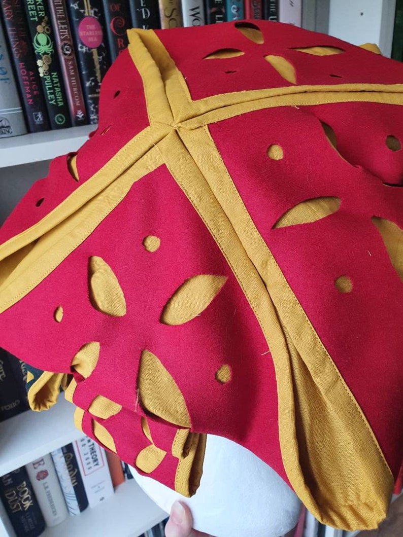 Puede incluir: Un sombrero de tela roja con ribete dorado y dise&ntilde;os recortados. El sombrero est&aacute; doblado y parece ser de fieltro.