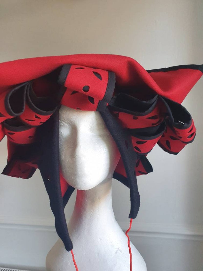 Puede incluir: Un sombrero de fieltro rojo con ribetes negros y dise&ntilde;os recortados. El sombrero tiene una ala ancha y est&aacute; decorado con m&uacute;ltiples capas de tela.