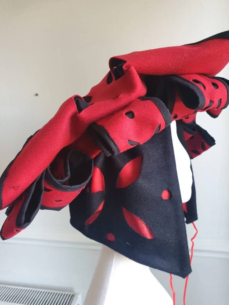 Puede incluir: Un sombrero de fieltro rojo y negro con dise&ntilde;os recortados. El sombrero est&aacute; doblado y tiene una larga cuerda roja atada.