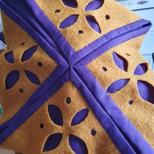 Puede incluir: Una tela de fieltro marr&oacute;n con ribete morado y dise&ntilde;os florales recortados. La tela est&aacute; doblada en forma de cuadrado.