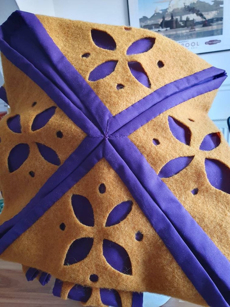 Puede incluir: Una tela de fieltro marr&oacute;n con ribete morado y dise&ntilde;os florales recortados. La tela est&aacute; doblada en forma de cuadrado.