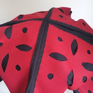 Puede incluir: Sombrero de fieltro rojo con recortes negros en forma de flores. El sombrero tiene un ribete negro alrededor de los bordes.