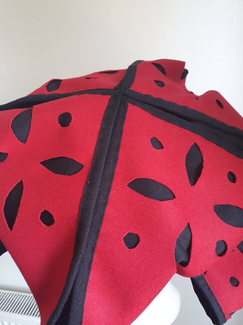 Puede incluir: Sombrero de fieltro rojo con recortes negros en forma de flores. El sombrero tiene un ribete negro alrededor de los bordes.
