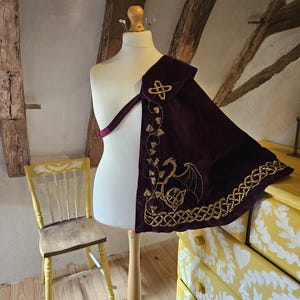 May include: A deep purple velvet cape draped over a mannequin, featuring gold embroidered dragon and vine motifs, a gold Celtic knot emblem at the shoulder, and a thick gold braided trim along the bottom edge, secured with a matching purple strap.