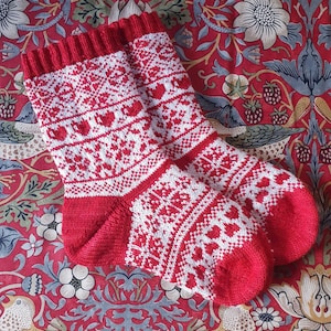 May include: A pair of red and white knitted socks with a heart pattern. The socks are on a red and white floral fabric.